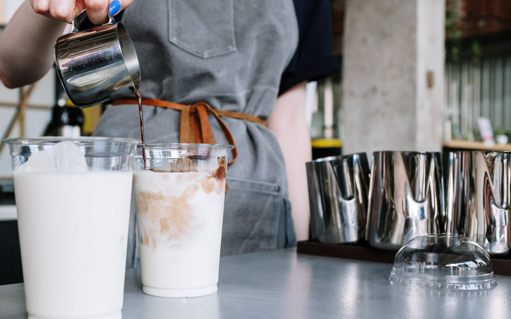 A barista pours espresso into cold milk in plastic cups.